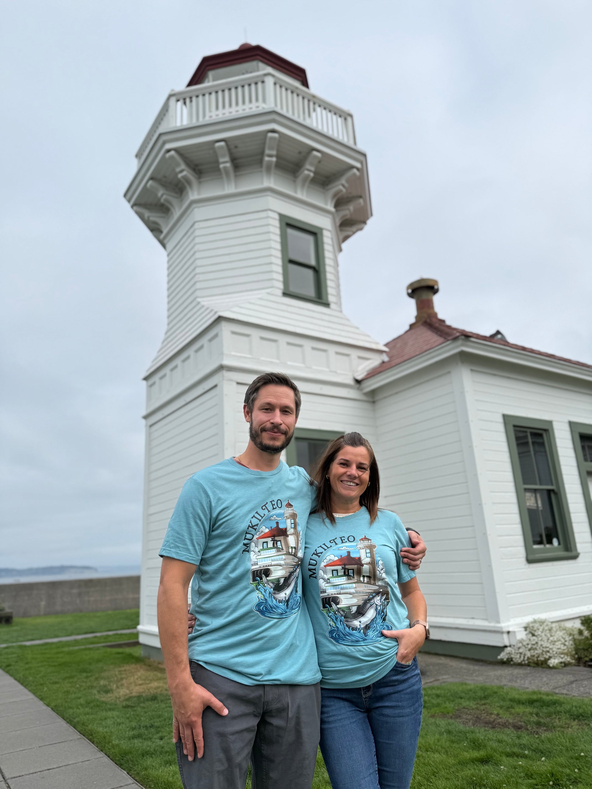 Two people wearing matching blue t-shirts with a lighthouse design in front of a white lighthouse.