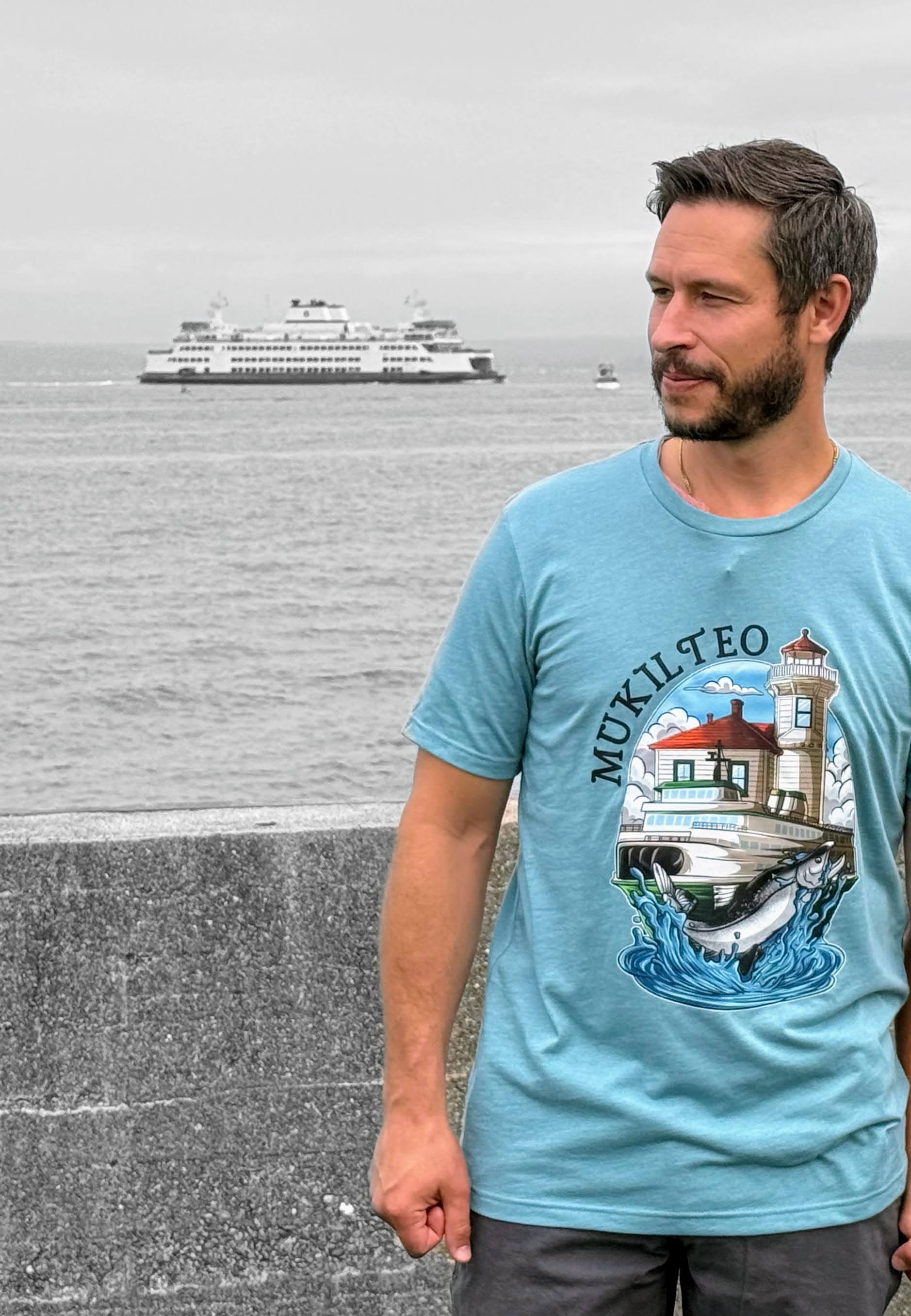 Man wearing a blue t-shirt with a lighthouse design, standing by a waterfront with a ferry in the background.
